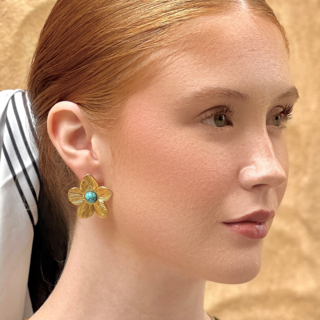 Model wearing gold floral stud earrings with turquoise stone center, styled with sleek hair and neutral makeup.